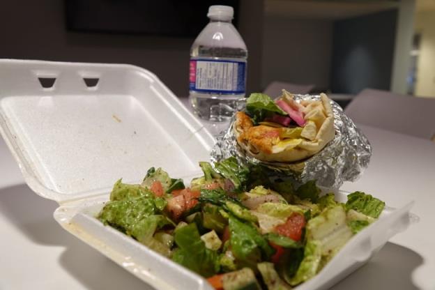 A salad covered in dressing is shown in a white Styrofoam co<em></em>ntainer with a bottle of water in the background and a wrap with pickles, turnip and chicken sticking out of it. 