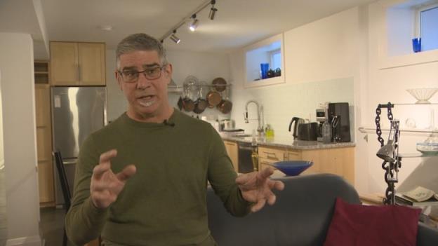 A man inside an apartment gestures as he speaks to the camera. 